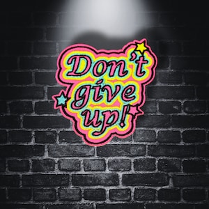 Könnte beinhalten: Ein gestickter Aufnäher in Pink, Gelb und Blau mit dem Text "Don't give up!" vor einem Hintergrund aus schwarzen Ziegeln.