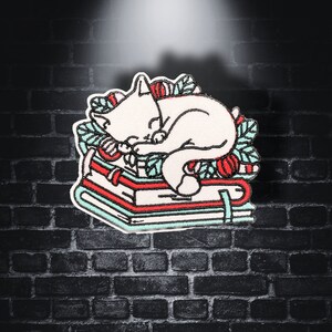 May include: White cat sleeping on a stack of three books with red and green floral designs. The cat is curled up on the top book, with its head resting on the second book. The books are stacked on top of each other, with the bottom book being the largest.