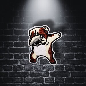 May include: Embroidered patch of a bulldog in a dabbing pose. The patch has a white background with brown and white details. The dog's arm is raised, and its face is partially covered. The patch is set against a dark brick wall.