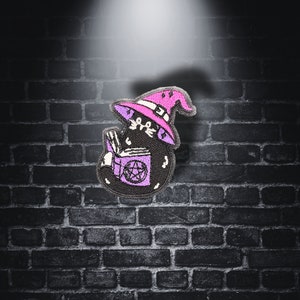 Könnte beinhalten: Eine schwarze Katze mit einem rosa Hexenhut und einem lila Buch mit einem Pentagramm auf dem Cover. Die Katze sitzt auf einer Backsteinmauer.