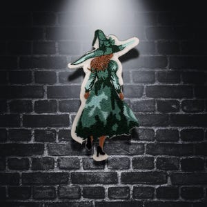 Peut inclure: Écusson brodé représentant une sorcière en robe et chapeau verts, vue de dos. La sorcière a les cheveux roux et porte des chaussures noires. L'écusson a une bordure blanche et est posé sur un mur de briques sombres.
