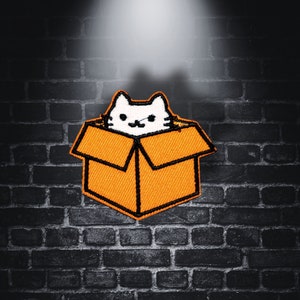 Puede incluir: Un gato blanco con ojos negros asoma de una caja de cartón naranja. La caja tiene una solapa en la parte superior y está sentada sobre un fondo de ladrillos oscuros.