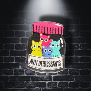 May include: A black and white embroidered patch of a glass jar with a pink lid. The jar is filled with four colorful cartoon cats. The text "Anti Depressants" is written on the front of the jar.