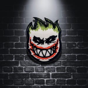 May include: Embroidered patch featuring a stylized, grinning face with green flame-like hair, black eyes, and a red mouth with white teeth. The patch is set against a dark brick wall background.