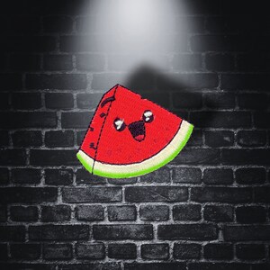 May include: A red watermelon slice with a smiling face embroidered on it. The watermelon is against a dark brick background.
