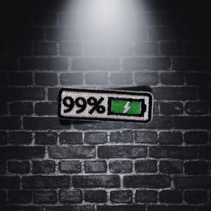 Könnte beinhalten: Ein gestickter Patch in Schwarzweiß mit einem Batteriesymbol und dem Text "99%" auf einem Hintergrund aus Ziegeln.