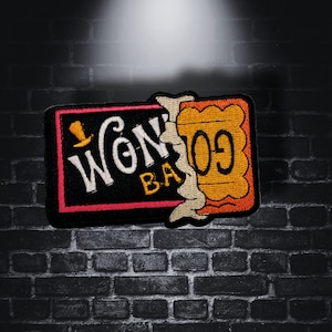 Könnte beinhalten: Ein schwarzer und rosa gestickter Patch mit dem Text "Wonka Bar" und einem Schokoladenriegel-Design. Der Patch befindet sich auf einem Hintergrund aus Ziegelmauer.