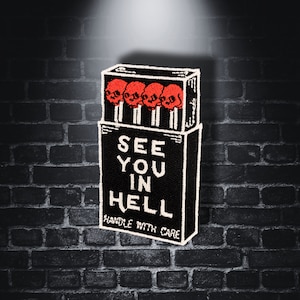 Op de afbeelding: Zwarte en witte geborduurde patch met een doosje lucifers. De lucifers hebben rode schedelkoppen en het doosje zegt "See You In Hell" met de tekst "Handle With Care" onderaan.