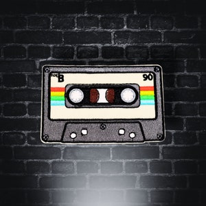 Könnte beinhalten: Ein Kassettenband im Vintage-Stil mit einem schwarzen und grauen Kunststoffgehäuse. Das Band ist mit "90" beschriftet und hat ein Regenbogenstreifen-Design.