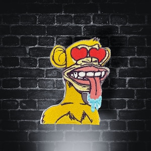 May include: Embroidered patch of a yellow monkey with red heart-shaped eyes and a long tongue. The monkey's mouth is open, showing teeth and a pink tongue. Blue drool drips from the tongue. Set against a dark brick wall.