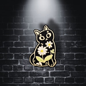 Pode incluir: Patch de gato preto bordado com flores brancas e folhas verdes. O gato está sentado com a cabeça inclinada para o lado.