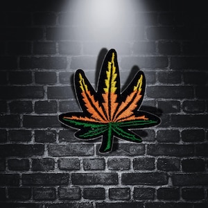 May include: A green, orange, and yellow embroidered marijuana leaf patch. The patch is on a black brick background.
