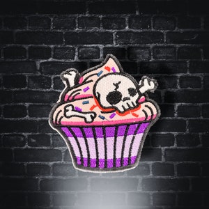 Può includere: Un cupcake rosa e viola con un teschio e ossa incrociate in cima. Il cupcake è decorato con glassa rosa e codette colorate.