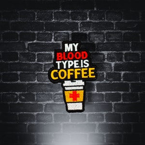 May include: A black brick wall with a colorful embroidered patch that says "My blood type is coffee" with a red cross on a yellow coffee cup.