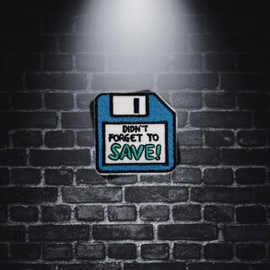 Può includere: Un floppy disk blu con un'etichetta bianca su cui è scritto "Didn't forget to SAVE!" in testo verde. Il floppy disk è su un muro di mattoni scuri.