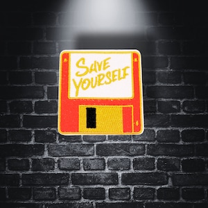 May include: A red and yellow floppy disk with the text "Save Yourself" embroidered on it. The floppy disk is against a dark brick background.