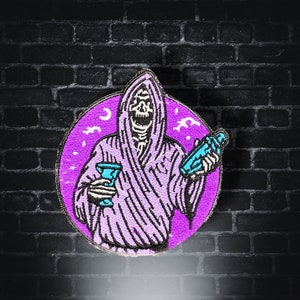 Op de afbeelding: Geborduurde patch met een Grim Reaper-figuur in een paarse mantel, met een cocktailshaker en een glas. Het ontwerp staat tegen een paarse cirkel met een maan en sterren, met een zwarte bakstenen achtergrond.