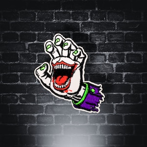 May include: Embroidered patch featuring a stylized hand with a wide, open mouth and sharp teeth. The hand has green fingernails and is wearing a purple sleeve with a green trim. The patch is set against a dark brick wall.