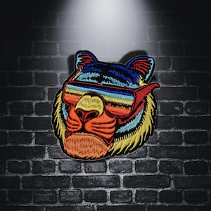Pode incluir: Patch bordado com a cabeça de um tigre usando óculos de sol. O patch tem um esquema de cores vibrantes, com a pelagem do tigre em amarelo, azul e laranja. Os óculos de sol têm uma lente colorida em arco-íris. Para roupas ou acessórios.