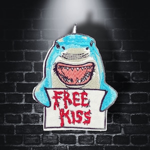 Può includere: Uno squalo blu con un ghigno pieno di denti tiene un cartello bianco che dice "FREE KISS" in lettere rosse. Lo squalo è ricamato e ha un effetto 3D.
