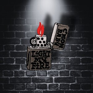 Pode incluir: Um remendo bordado em cinza e preto que representa um isqueiro com o texto "Light My Fire" na frente. O isqueiro está fechado, com a chama visível.