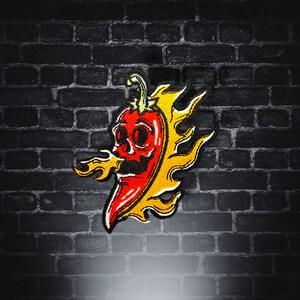 Puede incluir: Un parche bordado con un chile rojo con una cara de calavera y llamas, en amarillo y rojo.