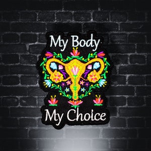 Op de afbeelding: Geborduurde patch met de tekst "My Body My Choice". De patch heeft een kleurrijk bloemmotief rond een gestileerde baarmoeder in geel, roze en blauw. De patch staat op een zwarte achtergrond.