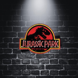 Puede incluir: Parche bordado con el logotipo de Jurassic Park. El diseño incluye un círculo rojo con una silueta de dinosaurio negro, un borde amarillo y las palabras "JURASSIC PARK" en amarillo y blanco. Forma rectangular.