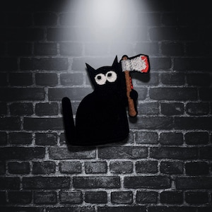 Puede incluir: Parche de gato negro bordado con grandes ojos blancos, sosteniendo un pequeño hacha con una hoja roja y blanca. El gato tiene cola. Sobre un fondo de pared de ladrillo oscuro.