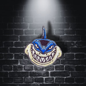 Può includere: Una toppa di squalo ricamata in blu e bianco con un ghigno pieno di denti. Lo squalo ha un corpo blu, denti bianchi e uno sfondo giallo.