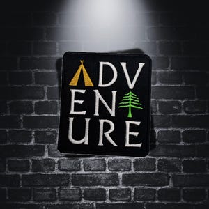 Può includere: Toppa quadrata nera con la scritta "ADVENTURE" ricamata in bianco. La lettera "A" presenta un teepee giallo e la lettera "E" un pino verde. La toppa è contro un muro di mattoni scuri.