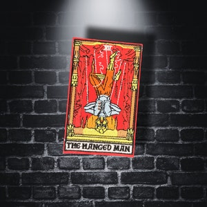 Op de afbeelding: Een geborduurde patch met de tarotkaart De Gehangene. De figuur hangt ondersteboven met een halo en een touw om zijn enkel. De kaart heeft de tekst "De Gehangene".