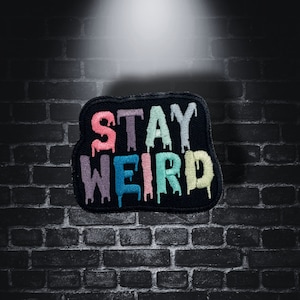 Op de afbeelding: Een zwart geborduurd embleem met de woorden "STAY WEIRD" in kleurrijke, druipende letters. De patch heeft een unieke, onregelmatige vorm en staat tegen een donkere bakstenen muur.