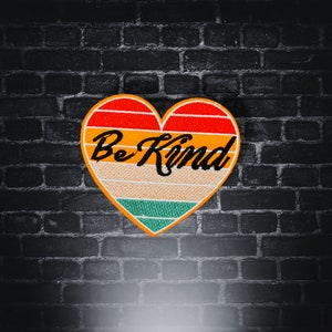 Op de afbeelding: Geborduurde hartvormige patch met de woorden "Be Kind" in zwart schrift. De patch heeft een retro kleurenschema met rode, oranje, gele, groene en witte strepen. De patch staat tegen een donkere bakstenen muur.