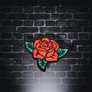 Peut inclure: Une rose rouge brodée avec des feuilles vertes sur un fond de briques noires.