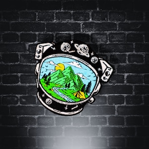 Op de afbeelding: Geborduurde patch met een astronautenhelm ontwerp. Het vizier van de helm toont een landschap met bergen, een rivier, bomen en een tent onder een gele zon. De patch is zwart met witte en kleurrijke accenten, ideaal voor jassen of tassen.