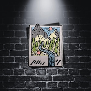 Peut inclure: Un patch brodé représentant un paysage de montagne avec une rivière qui le traverse. Le patch est dans un cadre blanc avec une bordure de style Polaroid et le texte "#1111" en bas.