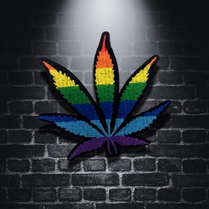 Op de afbeelding: Geborduurde cannabisblad patch met een regenboogkleurenschema. De patch heeft een zwarte omtrek en is geplaatst tegen een donkere bakstenen muur achtergrond. Het bladontwerp is gevuld met levendige kleuren, waaronder rood, oranje, geel, groen, blauw en paars.