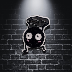 May include: Embroidered patch featuring a black, round character with large white eyes, pink cheeks, and a chef's hat. The patch has a running pose and is set against a dark brick wall.