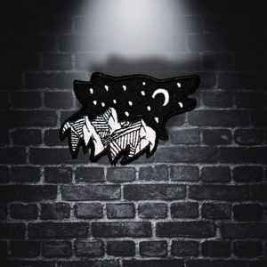 May include: Black and white embroidered patch of a wolf howling at the moon with a mountain range behind it.