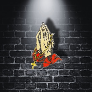 Peut inclure: Un patch brodé rouge et jaune représentant une paire de mains jointes en prière avec une chaîne dorée et une croix.