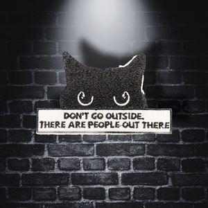 May include: A black cat peeking out from behind a brick wall with a white sign that reads "Don't go outside. There are people out there."