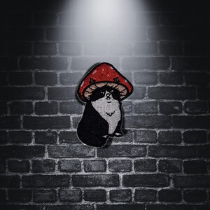 Può includere: Toppa ricamata raffigurante un gatto bianco e nero con un cappello a fungo rosso. La toppa presenta un design dettagliato con cuciture visibili. Lo sfondo è un muro di mattoni scuri.
