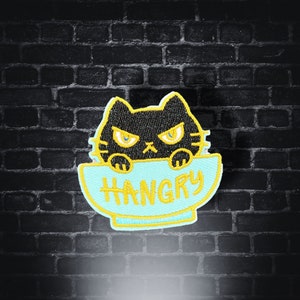 May include: Black cat with yellow eyes peeking over the rim of a blue bowl with the word "HANGRY" written in yellow letters.