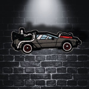 Peut inclure: Un patch brodé représentant une DeLorean, la machine à voyager dans le temps du film Retour vers le futur. La voiture est grise et noire avec une bande rouge et blanche à l'arrière et un tuyau d'échappement argenté.
