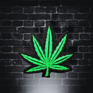May include: A green marijuana leaf patch with black stitching, against a dark brick wall background.