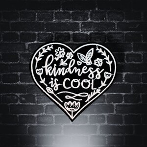 May include: Black heart-shaped patch with white embroidery. The patch features the text "kindness is cool" with floral designs around the edges.