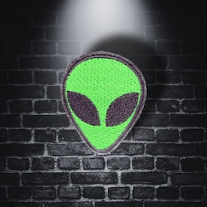 May include: A green alien face embroidered patch with black eyes. The patch is on a dark brick background.