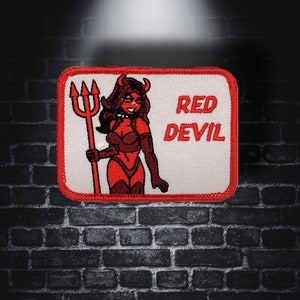 Peut inclure: Patch "Red Devil" avec un fond blanc et des coutures rouges. Le patch présente une image de dessin animé d'une femme aux cheveux roux avec des cornes et une queue, tenant une fourche. Le texte "RED DEVIL" est imprimé en rouge.