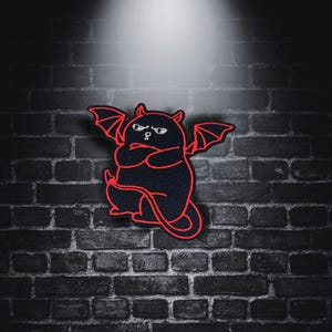 May include: A black and red embroidered patch of a cartoon devil cat with wings and a tail. The cat has a grumpy expression and is standing with its arms crossed.